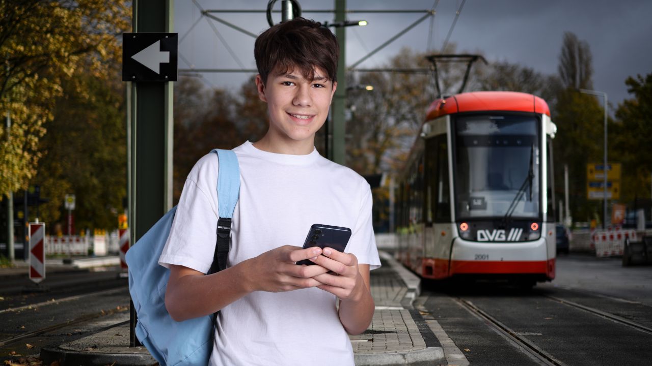 Junger Mann mit Smartphone an einer DVG-Haltestelle mit einfahrender Bahn