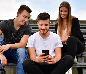 zwei junge Männer und eine junge Frau sitzen auf Stufen und schauen in ein Smartphone