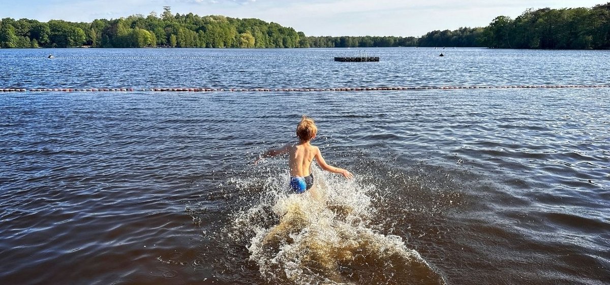 Ein Kind läuft an einem sonnigen Tag vom Sandstrand ins Wasser des Freibads Wolfssee. Im Hintergrund sind der See, eine Badeplattform und bewaldete Ufer zu sehen.