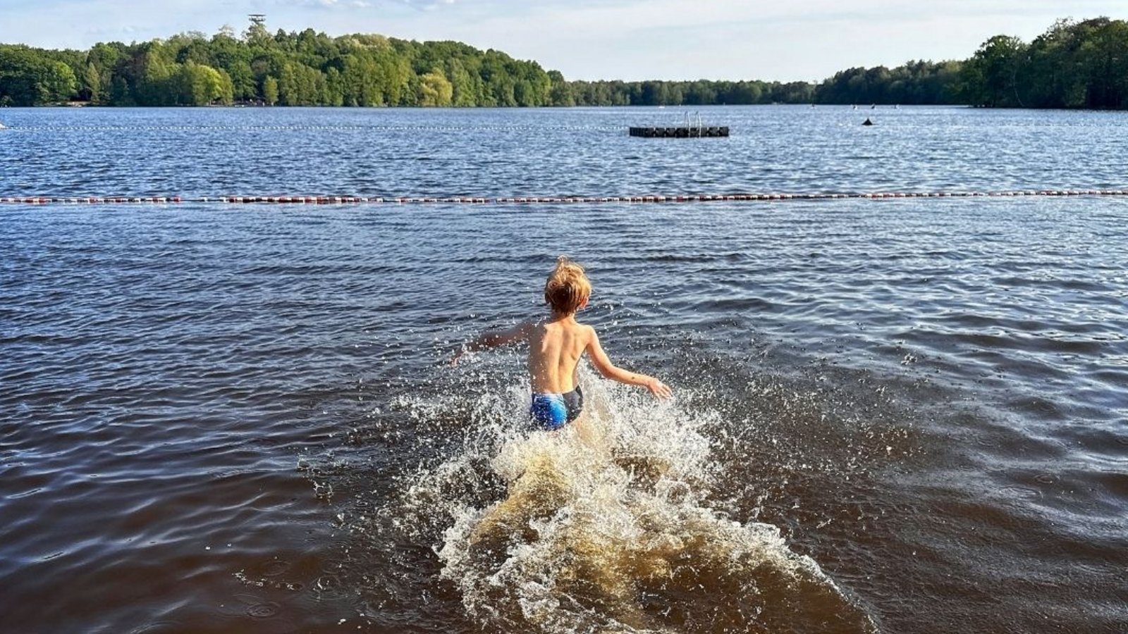 Ein Kind läuft an einem sonnigen Tag vom Sandstrand ins Wasser des Freibads Wolfssee. Im Hintergrund sind der See, eine Badeplattform und bewaldete Ufer zu sehen.